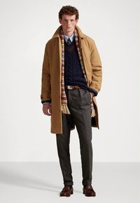 Bruine overjas, marineblauwe kabelgebreide trui, sjaal met patroon, grijze broek en donkere schoenen. Gestructureerd ontwerp met een getailleerde pasvorm.