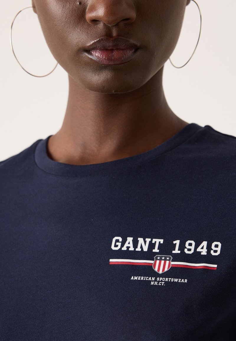 Granatowa bawełniana koszulka z krótkim rękawem z białym napisem "GANT 1949" oraz logo, okrągły dekolt, gładka tekstura i casualowy design.