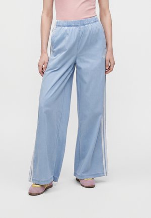 Person, die hellblaue, weit geschnittene Hose mit weißen Seitenstreifen, ein pinkes Oberteil und violette Sneakers trägt, steht vor einem einfarbigen hellen Hintergrund.