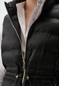 Schwarze Pufferrweste mit hohem Kragen, Reißverschluss vorne, elastischem Kordelzug in der Taille und glänzendem silbernen Reißverschlussdetails.