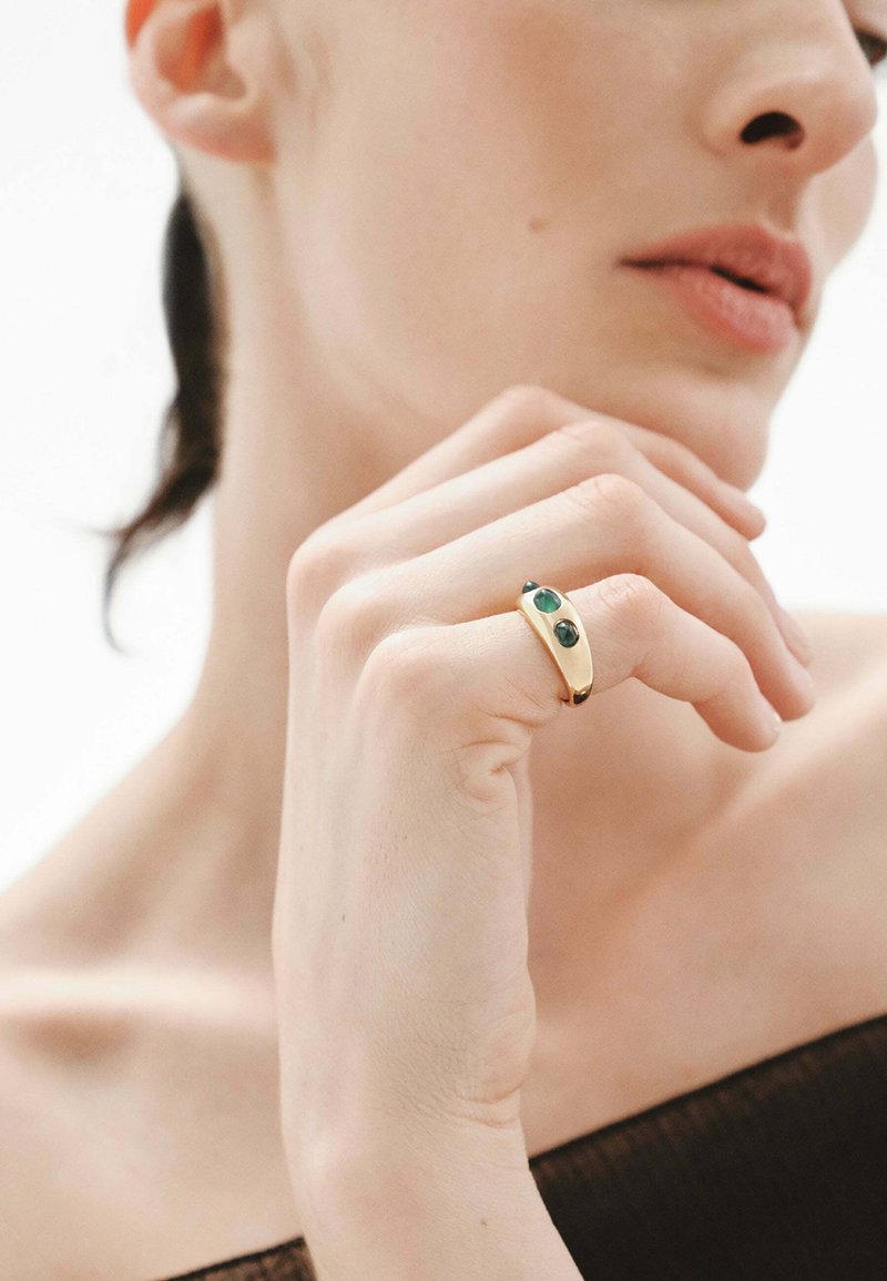 Anillo de oro con dos piedras verdes; diseño curvado; textura suave; llevado en un dedo; la mano del modelo está posicionada cerca de la cara con un fondo neutro.