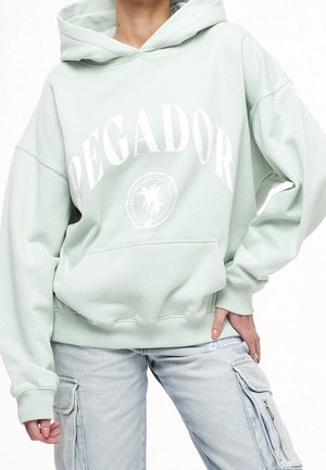 Person wearing light green oversized hoodie with "PEGADOR" text and palm tree graphic, paired with light-wash cargo jeans.