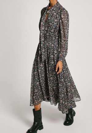 Kobieta w długiej, prześwitującej sukience w kwiaty z bufkami na rękawach, do której dobrała czarne skórzane botki do połowy łydki z klamrami.