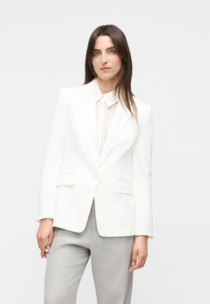 Mujer con cabello largo y castaño, vestida con un blazer blanco y pantalones gris claro, de pie contra un fondo claro y uniforme.