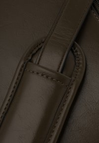 Szczegół brązowej torby skórzanej, pokazujący teksturowaną powierzchnię, gładki pasek i kontrastujące przeszycia. Posiada zaokrąglone krawędzie i ogólny subtelny design.