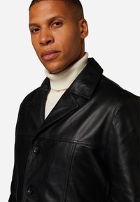 Schwarze Lederjacke mit Knöpfen, breitem Kragen und glatter Textur, getragen über einem cremefarbenen Rollkragenpullover. Hat einen klassischen Stil und eine taillierte Passform.