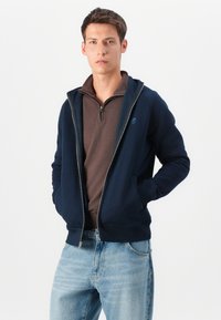 Hanorac cu fermoar de culoare bleumarin, cu un design monochrom, mâneci raglan și buzunare frontale, purtat peste o cămașă cu fermoar de culoare maronie. Jeans casual din denim.