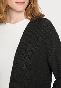 Cardigan nero a maglia con un design aperto sul fronte, caratterizzato da una texture a costine e maniche lunghe, indossato sopra una camicia bianca.