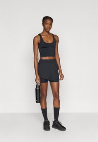 Conjunto deportivo negro de top corto y shorts, con una tela texturizada. Botella deportiva negra en la mano derecha, completan el look calcetines y zapatillas negras.