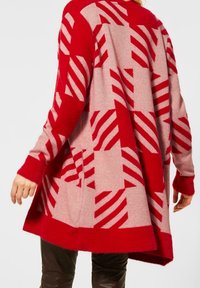 Frau trägt eine pink-rote, übergroße Strickjacke mit geometrischen Streifenmustern, kombiniert mit dunkler Hose, von hinten gezeigt.