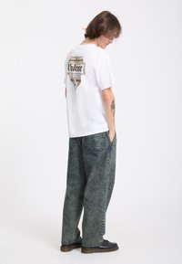 Jeune personne aux cheveux mi-longs porte un t-shirt blanc imprimé, un jean large délavé et des chaussures noires, debout les mains dans les poches.