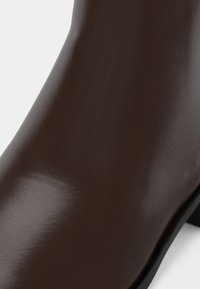 Gros plan sur une botte en cuir brun foncé lisse montrant le dessus incurvé ainsi qu'une partie de la semelle noire et du talon bloc.