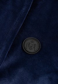 Laivastonsininen ribbineulospaita, jossa pystysuoria raitoja ja pyöreä musta kumilogo. Materiaali on pehmeää ja sileäpintaista.