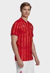 Camiseta deportiva roja con rayas verticales, de manga corta y cuello en V. Presenta detalles del logo en el pecho y el hombro. Tejido texturizado.