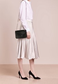 Donna in blusa bianca, gonna midi plissettata argento, tacchi alti neri e borsa a tracolla con catena nera in piedi contro uno sfondo neutro.