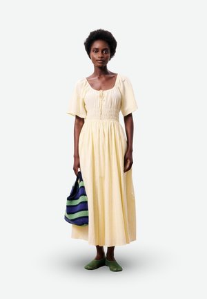 Femme portant une robe jaune pâle à manches courtes et longueur cheville, tenant un sac rayé bleu et vert et chaussant des chaussures vertes sans lacets.