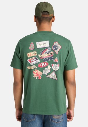 T-shirt verde in cotone con vari pezzi di tessuto colorati sul retro, tra cui temi naturalistici, loghi e un disegno di una cassetta.