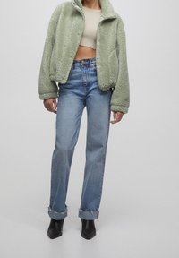 Pelsagt grøn cropped jakke med høj krave, parret med lyseblå højtaljede jeans og sorte hælede ankelstøvler.