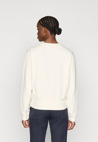 Sudadera cremosa con cuello redondo, puños y dobladillo acanalados. Textura suave, corte relajado y vista desde la parte posterior contra un fondo liso.
