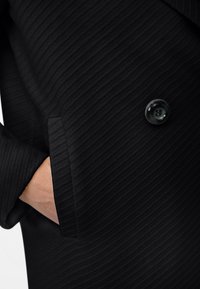 Tessuto nero testurizzato con un design a righe, dettaglio con bottone sulla manica e tasca laterale. Sartoria semplice ed elegante in una silhouette aderente.
