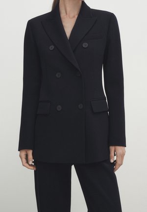 Femme portant un blazer noir cintré à double boutonnage avec des revers crantés, des poches à rabat, et un pantalon noir assorti sur un fond uni.