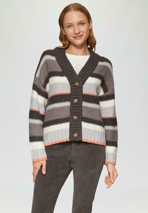Femme portant un cardigan rayé boutonné avec des nuances de gris, blanc et pêche, associé à un pantalon gris foncé, debout devant un fond uni.