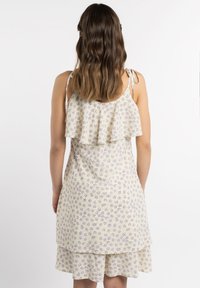 Rochie de soare florală în galben pal cu flori mov. Prezintă un overlay cu volane, bretele reglabile și o margine în straturi. Textură de țesătură moale.