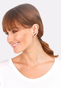 Drei schwarze Steckerohrringe in unterschiedlichen Größen, im Ohrläppchen eines Models gesteckt. Das Model hat glattes braunes Haar und trägt ein weißes Oberteil.
