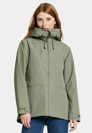 Frau mit roten Haaren, die eine hellgrüne wasserdichte Kapuzenjacke und blaue Jeans trägt, steht vor einem schlichten Hintergrund.