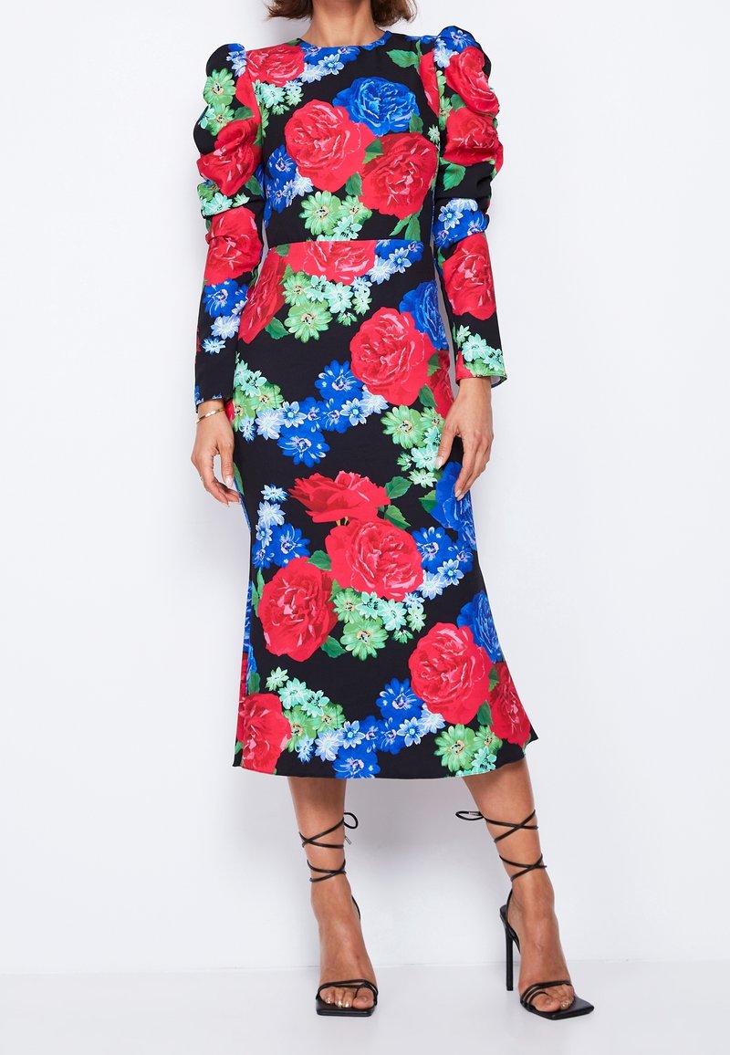Femme portant une robe midi noire avec de grands motifs floraux rouges, bleus et verts et des manches longues bouffantes, associée à des sandales noires à talons hauts.