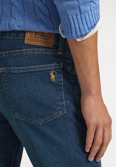 Calças de ganga azul-escuro com acabamento texturizado, apresentando uma etiqueta da marca em couro e um pequeno logótipo bordado de cavalo no bolso traseiro.