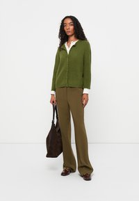 Olivgrön ribbad cardigan med knappar, i kombination med olivfärgade vida byxor. En vit skjorta med krage under; brun mockaväska och skor.