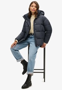 Marine pufferjack met een capuchon, knoopsluiting en doorgestikt textuur. Gecombineerd met lichtblauwe, opgerolde jeans en zwarte veterschoenen.