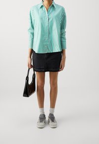 Grün-weißer, vertikal gestreifter Hemd, schwarze Denim-Rock mit Fransen, schwarze Handtasche, graue Sneakers mit weißen Sohlen und Socken.