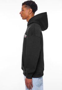 Sweatshirt à capuche noire avec une poche kangourou, comportant un texte brodé sur la poitrine. Tissu doux et détails de poignets côtelés.