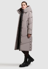 Lang, polstret frakk i lys beige med høj krave. Har sorte detaljer, trykknapper og sidelommer med lynlås. Tekstureret, quiltet design.