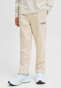 Beige joggings met zijzakken en een zwart logo op de linken bovenbeen, gecombineerd met witte sportschoenen met een gestructureerde bovenkant en een dikke zool.