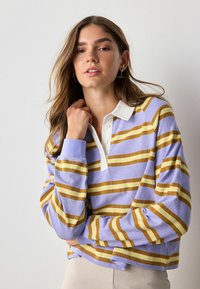 Giovane donna con i capelli castano chiaro sciolti, che indossa un maglione a strisce lavanda, giallo e bianco con colletto bianco, in posa davanti a uno sfondo neutro.