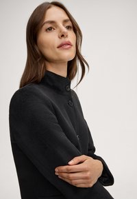 Zwarte wollen jas met een hoge kraag, voorzien van gestructureerde schouders, twee zwarte knopen en een gestructureerd oppervlak. Armen gekruist op de borst.