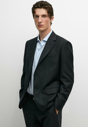 Jonge man met krullend haar draagt een zwart colbert en lichtblauw overhemd, staat met handen in de zakken tegen een effen witte achtergrond.