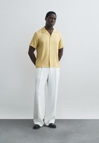 Άνδρας που στέκεται ανάμεσα σε λευκό τοίχο φορώντας κίτρινο πουκάμισο με κοντά μανίκια και κουμπιά, φαρδιά λευκά παντελόνια και μαύρα παπούτσια.