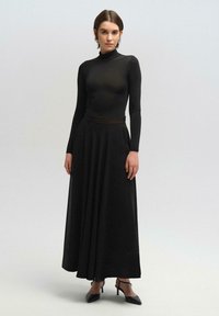Vestido negro de manga larga con un corsé ajustado y falda acampanada. Elaborado en una tela suave, cuenta con un escote alto y un sutil detalle en la cintura, combinado con tacones negros.
