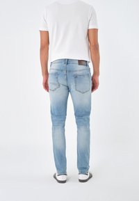 Pantalones vaqueros de denim azul claro con un corte ajustado, que cuentan con bolsillos traseros, un ligero desgastado y un parche de cuero en la cinturilla. Se lleva una camiseta blanca y zapatillas.
