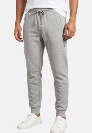 Hombre con pantalones jogger gris claro con cintura ajustable y zapatillas blancas, de pie frente a un fondo liso.