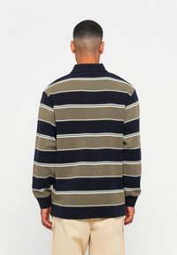 Man met kort haar in een langarmig navy en olijfgroene gestreepte shirt en beige broek, met zijn rug naar de camera tegen een effen achtergrond.