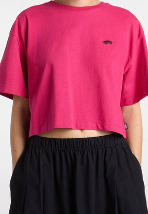 Person trägt ein kurz geschnittenes, knallpinkes T-Shirt mit kurzen Ärmeln und einem kleinen schwarzen gestickten Logo, kombiniert mit schwarzen Hosen mit elastischem Bund.