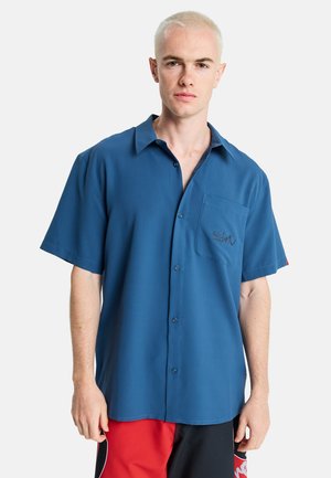 Koszula na guziki z krótkim rękawem z niebieskiej tkaniny, z kieszenią na lewej piersi i subtelnym logo. Gładka tekstura, luźny krój.