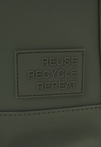 Un écusson texturé vert présente le texte en relief "REUTILISER RECYCLER RÉPÉTER" avec une bordure rectangulaire. Un détail de couture délimite l'écusson.
