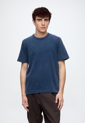 Jeune homme aux cheveux bouclés et foncés porte un t-shirt bleu foncé uni à manches courtes et un pantalon marron foncé sur un fond gris clair.