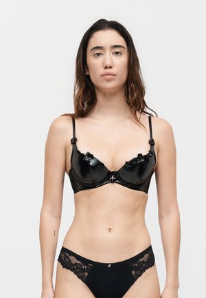 Femme aux longs cheveux bruns portant un soutien-gorge noir brillant avec des détails floraux et une culotte en dentelle noire, debout devant un fond clair uni.